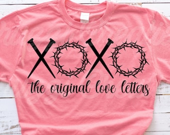 May include: A pink t-shirt with a black graphic design featuring a crown of thorns and nails forming the letters "XOXO" with the text "the original love letters" below.