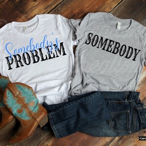 Könnte beinhalten: Zwei T-Shirts, eines weiß und eines grau, mit dem Text "Somebody's Problem" und "Somebody" bedruckt. Die Shirts liegen auf einer Holzoberfläche mit einer blauen Jeans und braunen Cowboystiefeln.