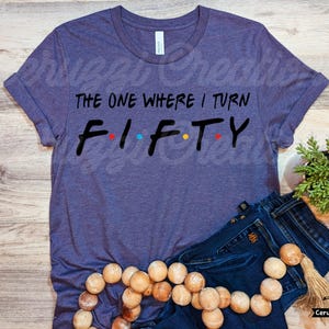 Może przedstawiać: Fioletowy T-shirt z napisem "The One Where I Turn Fifty" w kolorze czarnym z kolorowymi kropkami. Koszulka leży na drewnianej powierzchni z niebieskimi dżinsami i drewnianym naszyjnikiem z koralik&oacute;w.