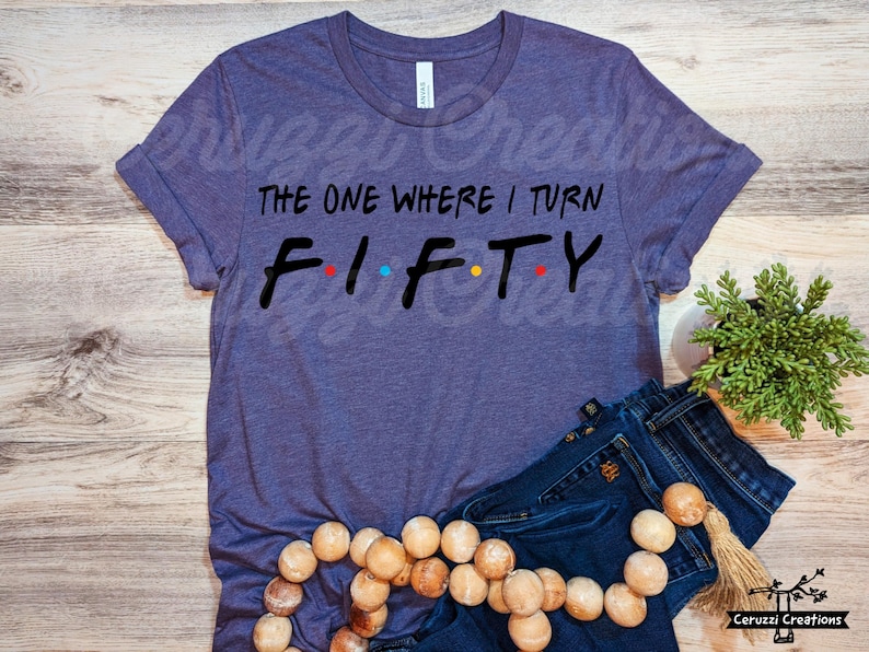 Może przedstawiać: Fioletowy T-shirt z napisem "The One Where I Turn Fifty" w kolorze czarnym z kolorowymi kropkami. Koszulka leży na drewnianej powierzchni z niebieskimi dżinsami i drewnianym naszyjnikiem z koralik&oacute;w.