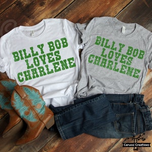 May include: Two t-shirts with the text "Billy Bob Loves Charlene" in green letters on a white and gray background. The shirts are laid out on a wooden surface with a pair of brown cowboy boots.