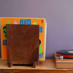 May include: A wooden record stand with a record leaning against it. The record cover is orange, green, and blue with the text "Elvis" on it. A turntable is on the table to the right of the stand.