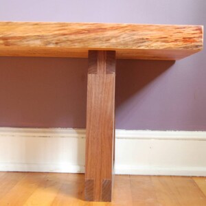 May include: A wooden bench with a natural wood finish and a single leg support. The bench is made of two different types of wood, one light and one dark. The bench is sitting on a wooden floor.