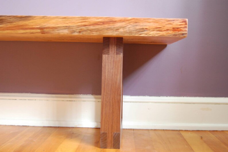 May include: A wooden bench with a natural wood finish and a single leg support. The bench is made of two different types of wood, one light and one dark. The bench is sitting on a wooden floor.