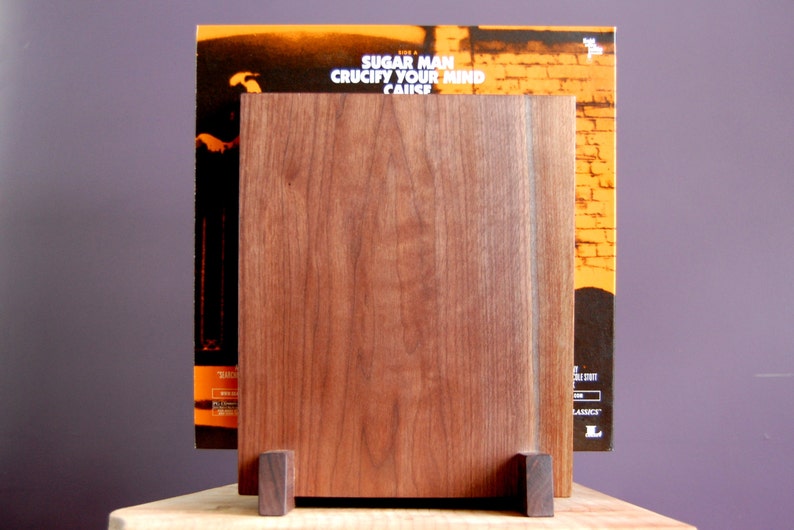 May include: A walnut wood record stand with a record leaning against it. The record cover is orange and features the text "Sugar Man Crucify Your Mind" in white.