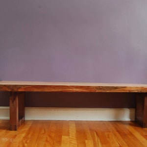 May include: A wooden bench with a natural wood finish and a simple design. The bench has a rectangular top and two legs on each side. The bench is sitting on a wooden floor.