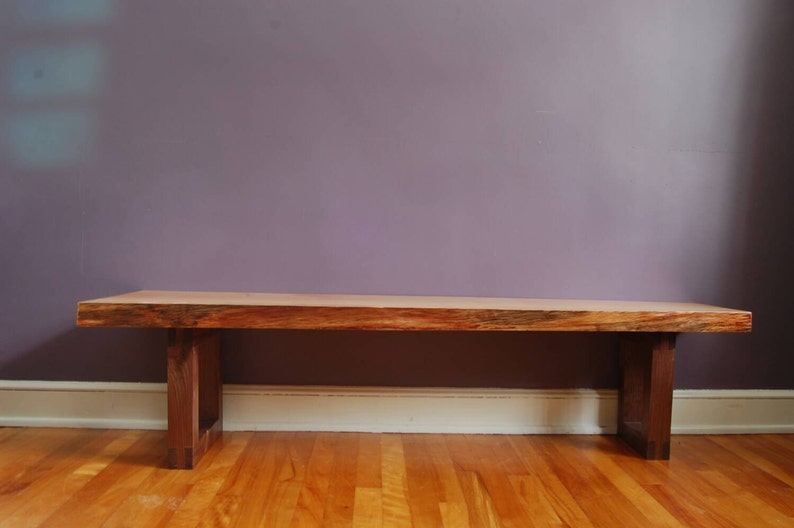 May include: A wooden bench with a natural wood finish and a simple design. The bench has a rectangular top and two legs on each side. The bench is sitting on a wooden floor.