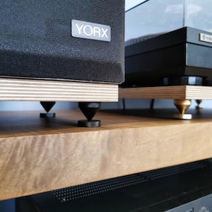 Peut inclure: Deux composants audio noirs, une platine vinyle et une enceinte, sont placés sur une étagère en bois. Les deux composants reposent sur des pieds coniques noirs. La platine vinyle porte l'inscription "KENWOOD". L'enceinte porte l'inscription "YORX".