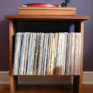 May include: A wooden record player stand with a turntable on top and a shelf below for storing vinyl records. The stand is made of dark wood and has a simple design.