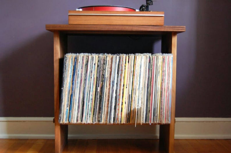 May include: A wooden record player stand with a turntable on top and a shelf below for storing vinyl records. The stand is made of dark wood and has a simple design.