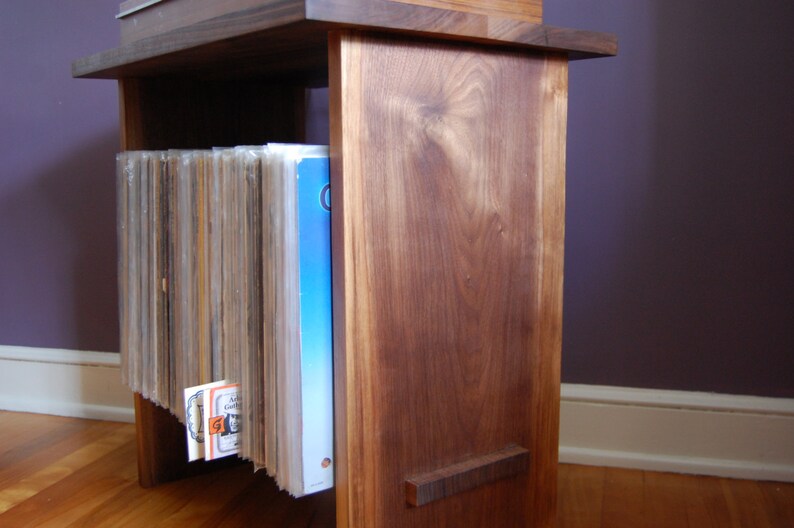 May include: A wooden record stand with a dark brown finish. The stand has a shelf for storing records and a vertical compartment for holding records upright. The stand is made of solid wood and has a simple, elegant design.