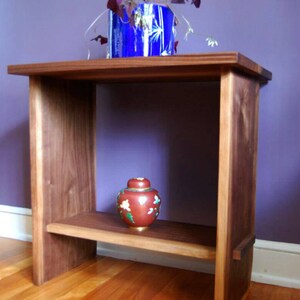 May include: A wooden end table with a single shelf. The table is made of dark wood and has a simple, modern design. A small, red and gold decorative jar sits on the shelf.
