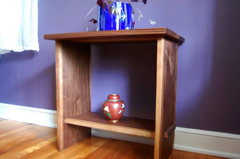 May include: A wooden end table with a single shelf. The table is made of dark wood and has a simple, modern design. A small, red and gold decorative jar sits on the shelf.