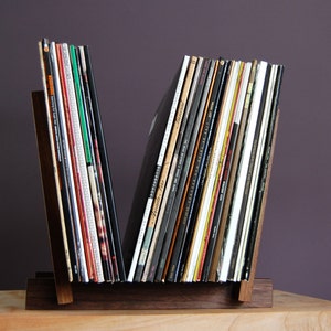 May include: A wooden record stand with a V-shaped design holds a collection of vinyl records. The stand is made of dark wood and has a natural finish. The records are arranged vertically and are mostly black with some colorful covers.