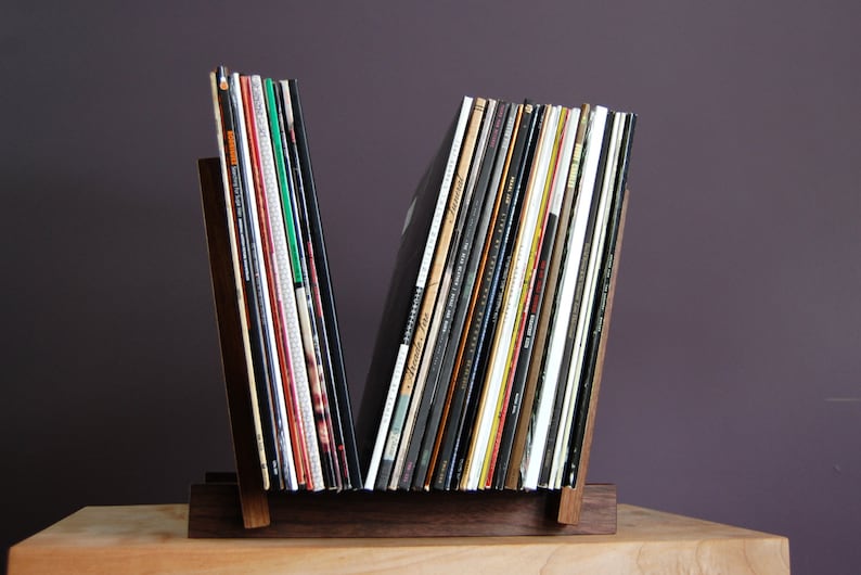 May include: A wooden record stand with a V-shaped design holds a collection of vinyl records. The stand is made of dark wood and has a natural finish. The records are arranged vertically and are mostly black with some colorful covers.
