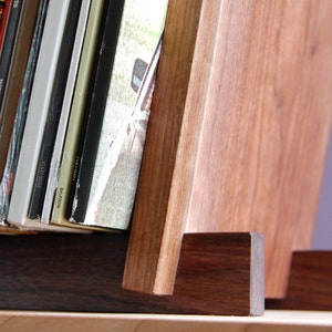 May include: A wooden record stand with a dark brown base and light brown upright supports. The stand is holding a stack of vinyl records.