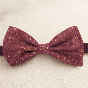 May include: A burgundy bow tie with a repeating gold diamond pattern. The bow tie is made of fabric and has a pre-tied bow shape. The background is a neutral, off-white color.