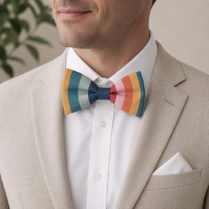 May include: A bow tie featuring vertical stripes in shades of mustard yellow, sage green, navy blue, pink, and orange. The bow tie is worn with a crisp white button-down shirt and a light beige blazer, with a white pocket square visible in the pocket.