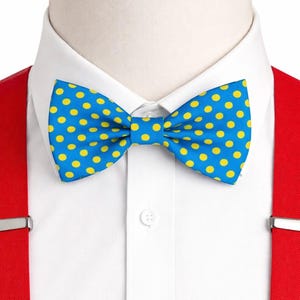 May include: A bright blue bow tie featuring a repeating pattern of small yellow polka dots, worn with a crisp white collared shirt and vibrant red suspenders with silver metal clips attached to the fabric on both sides of the shirt front.