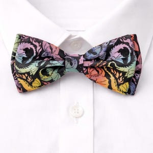 May include: A bow tie featuring a black background with a colorful, repeating pattern of stylized dragons in shades of yellow, orange, pink, purple, and blue. The bow tie is centered on a crisp white collared shirt with a single visible white button.