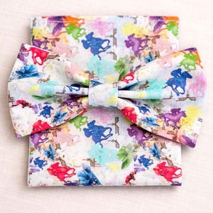 May include: A white fabric bow tie and matching pocket square set. The set features a colorful pattern of watercolor-style horses and jockeys in shades of blue, purple, red, and yellow. The bow tie is pre-tied.