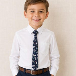 May include: A white button-down shirt with a navy blue tie featuring a pattern of video game controllers. The tie is worn with a brown belt and navy blue pants. The boy is smiling.