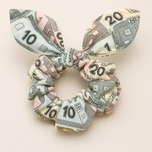 May include: A scrunchie with a bow, featuring a colorful Monopoly money print. The scrunchie is a mix of pastel colors, including light blue, pink, and yellow, with the numbers 1, 10, and 20 visible. The bow adds a playful touch.