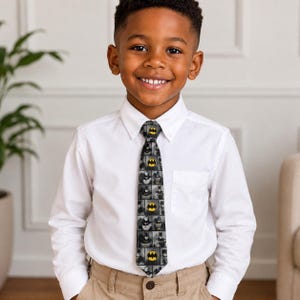 May include: A crisp white long-sleeved button-down shirt paired with a dark grey necktie featuring a repeating grid pattern of Batman comic book illustrations and the iconic yellow and black bat symbol logo, worn with khaki trousers.