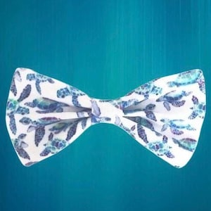 May include: A white bow tie featuring a pattern of blue and teal sea turtles. The bow tie is centered against a teal background, showcasing the detailed turtle design.