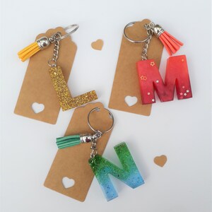 May include: Three resin letter keychains with glitter and a gradient colour effect. The letters are L, M, and N. Each keychain has a small tassel attached to the ring.