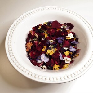 May include: A white ceramic bowl filled with a mix of colorful dried flower petals. The petals are primarily deep red, with accents of yellow, purple, orange, and white. The bowl has a decorative beaded rim.