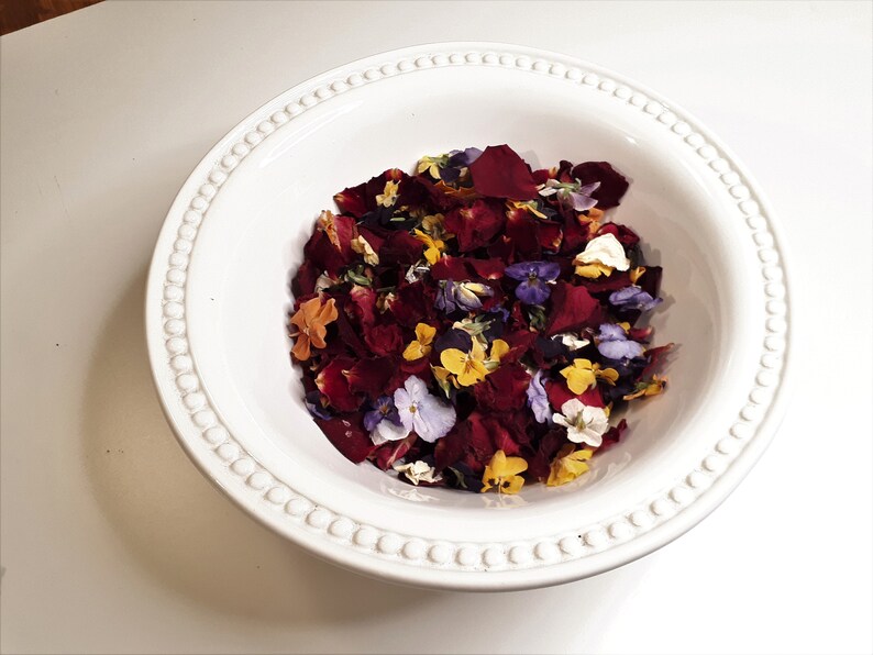 May include: A white ceramic bowl filled with a mix of colorful dried flower petals. The petals are primarily deep red, with accents of yellow, purple, orange, and white. The bowl has a decorative beaded rim.