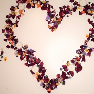 May include: A heart shape made of red rose petals and colorful flower petals, with four white tea light candles inside. The arrangement is on a white surface, creating a romantic and decorative display.