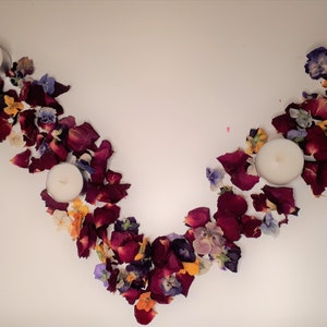 May include: An overhead shot of a decorative arrangement featuring three white candles and scattered flower petals. The petals are primarily deep red, with accents of yellow, purple, and white, creating a natural, organic aesthetic.