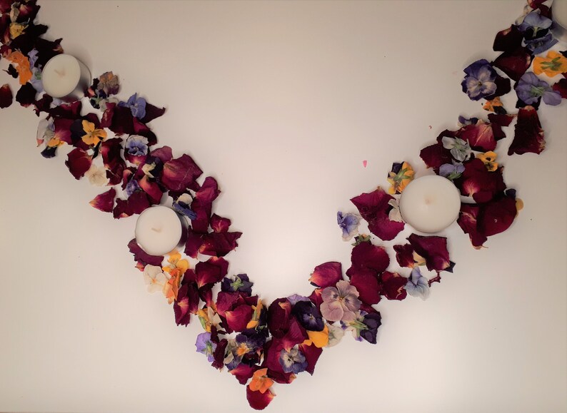 May include: An overhead shot of a decorative arrangement featuring three white candles and scattered flower petals. The petals are primarily deep red, with accents of yellow, purple, and white, creating a natural, organic aesthetic.