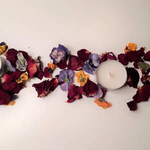 May include: Two white tea light candles are surrounded by dried rose petals and colorful flowers. The petals are a deep burgundy color, and the flowers are purple, yellow, and orange. The arrangement is on a white surface.