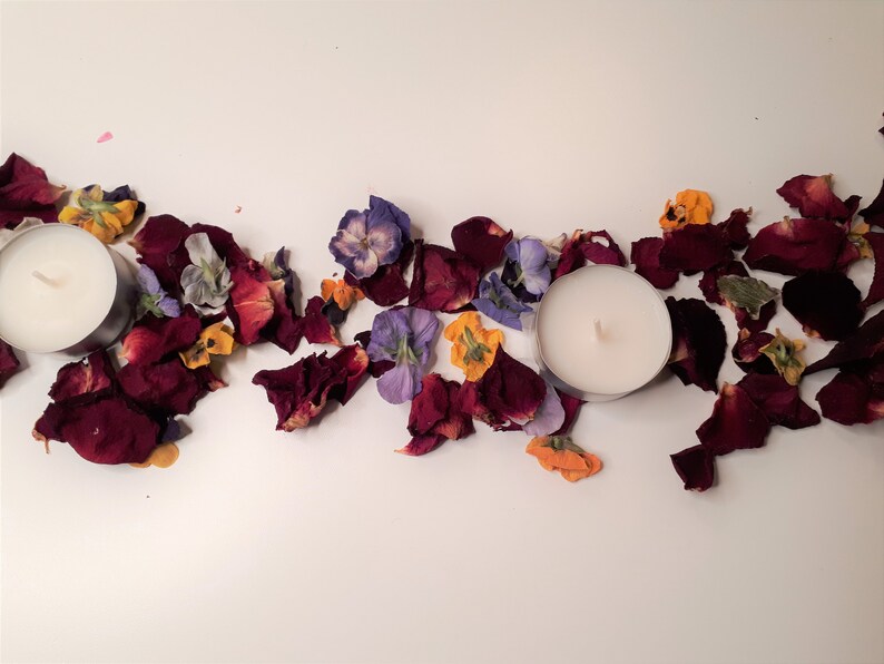 May include: Two white tea light candles are surrounded by dried rose petals and colorful flowers. The petals are a deep burgundy color, and the flowers are purple, yellow, and orange. The arrangement is on a white surface.