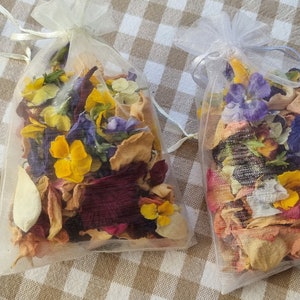 May include: Two small, sheer mesh bags filled with dried flowers and petals. The flowers are a mix of yellow, purple, and red, with beige and tan petals. The bags are tied with a white ribbon and sit on a tan and white checkered surface.