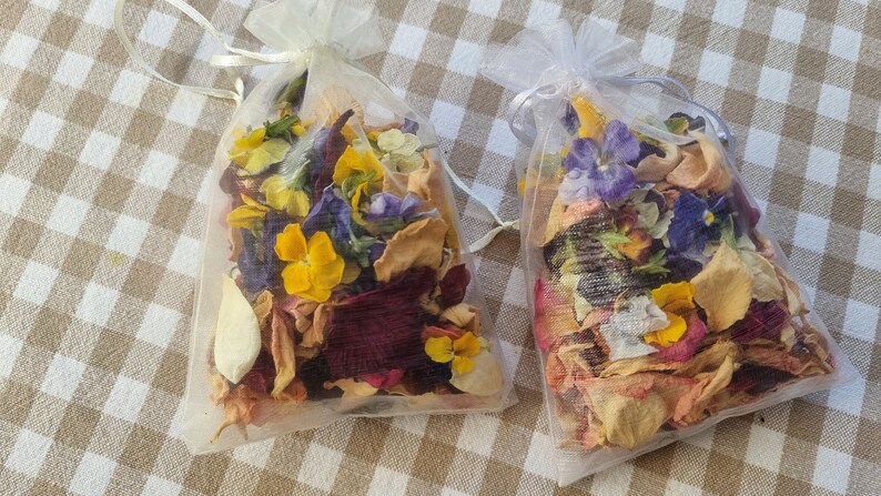May include: Two small, sheer mesh bags filled with dried flowers and petals. The flowers are a mix of yellow, purple, and red, with beige and tan petals. The bags are tied with a white ribbon and sit on a tan and white checkered surface.