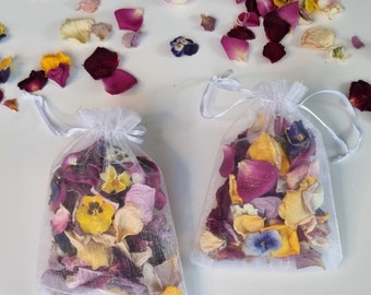 May include: Two small, white mesh bags filled with colorful dried flower petals and pansies. The petals are in shades of red, pink, yellow, and cream. Loose petals are scattered around the bags on a white surface. The bags have white ribbon ties.