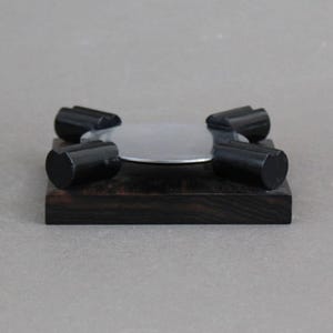 May include: A black wooden coaster with a silver metal top and four black knobs. The coaster is designed to hold a cup or glass.
