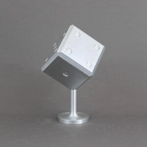 May include: A silver-coloured, oversized die sculpture. The die is mounted on a short, cylindrical stand. The die has six sides, each with a different number of circular indentations representing the numbers one to six.