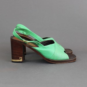 May include: A pair of green leather heeled sandals with a wooden platform and gold accents. The sandals have a slingback strap and an open toe.