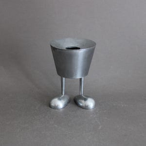 Peut inclure: Un petit récipient en métal argenté avec un corps conique et une ouverture circulaire sur le dessus. Le récipient a deux pieds cylindriques minces et deux pieds arrondis. L'objet est posé sur un fond gris.