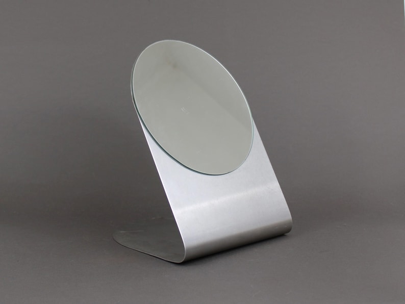 May include: A silver-coloured tabletop mirror with a round, slightly angled mirror surface.