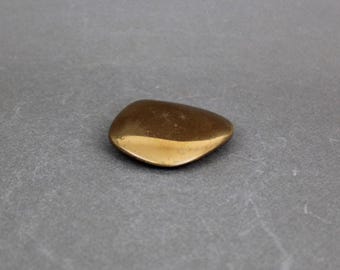 May include: A small, smooth, pebble-shaped object with a metallic appearance. The top surface is a darker, brownish-gold color, while the bottom half has a brighter, polished gold finish. The object is resting on a gray surface.