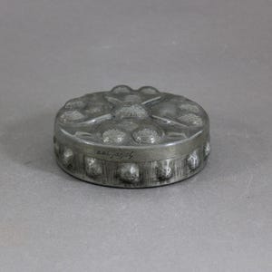 May include: A small, round, pewter-colored box with a decorative floral design on the lid. The box has a series of small, round embellishments around the base. The text "ael jagaf" is inscribed on the side.