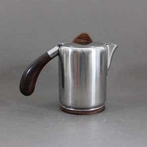 Può includere: Una caffettiera in acciaio inossidabile con manico, coperchio e base in legno scuro. La caffettiera ha un corpo cilindrico e un beccuccio per versare. Il manico è curvo verso l'esterno e il coperchio ha un piccolo pomello triangolare in legno.