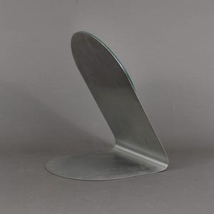 May include: A silver metal mirror stand with a curved design. The stand has a flat base and a curved back.