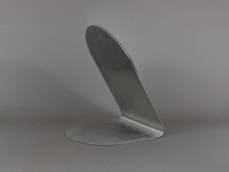 May include: A silver metal mirror stand with a curved design. The stand has a flat base and a curved back.